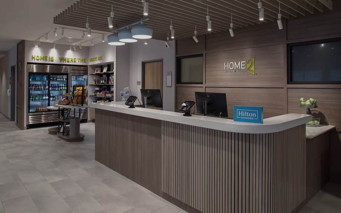 Front desk and market at the Home2 Suites Little Rock Midtown: slatted wood reception casework, a wood-slat ceiling with track lighting, and the retail coolers behind.