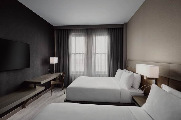 Double queen guestroom at the AC Hotel Kansas City Downtown: two queen beds with upholstered headboards, a wall-mounted television, a wood desk ledge with a table lamp, textured wall covering and sheer drapery.